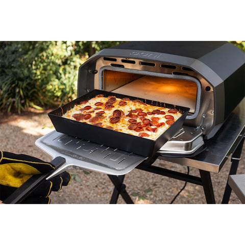 Eine Person mit Ofenhandschuhen holt mit dem Ooni Detroit-Style Pizzablech 32 x 24 cm eine frische Peperonipizza aus einem Outdoor-Pizzaofen, der auf einem Metalltisch aus Edelstahl steht. Im Hintergrund sind etwas Grün und eine Kiesfläche zu sehen.