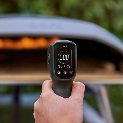 Une main tenant un thermomètre infrarouge numérique 2024 de Ooni indique une température de 500 degrés Fahrenheit. L'arrière-plan est légèrement flou et montre un four à pizza en plein air avec une flamme visible à l'intérieur, parfait pour réchauffer ces pierres à pizza.