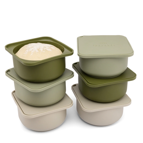 Una pila di cinque contenitori quadrati con coperchio. Tre contenitori e i coperchi sono di colore verde salvia, mentre due sono di colore beige chiaro. Il contenitore superiore della pila di sinistra contiene pasta lievitata cosparsa di farina. I coperchi sono stampati con la scritta "Babadoh", il che rende la confezione Babadoh ideale: 6 contenitori, scatola di olive e set di preparazione per pizza in legno di quercia.