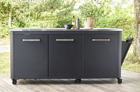 Die Cozze Aussenküche 180 mit Edelstahltischplatte steht auf einer Holzterrasse und besticht durch ihr schlichtes Design mit drei grauen Schränken für reichlich Stauraum. Eine Topfpflanze, eine Olivenölflasche und eine Schüssel verleihen dem Raum Charme, alles vor üppigem Grün im Hintergrund.