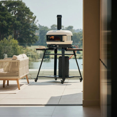 Ein Gozney Dome Gen 2 Pizzaofen - Hybrid Brennstoffsystem von Gozney wird auf einer Terrasse mit Glasgeländer und Blick auf Bäume und einen See präsentiert. Ein Korbstuhl neben dem Ofen sorgt für Komfort in diesem einladenden Außenbereich.