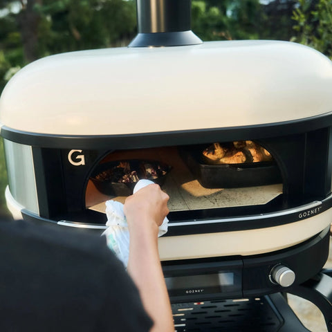Jemand benutzt den Gozney Dome Gen 2 Pizzaofen - Hybrid Brennstoffsystem von Gozney in seiner Outdoorküche und backt Essen in einer schwarzen Pfanne bei geöffneter Ofentür, während er ein Handtuch in der Hand hält. Dahinter ist verschwommenes Grün zu sehen.