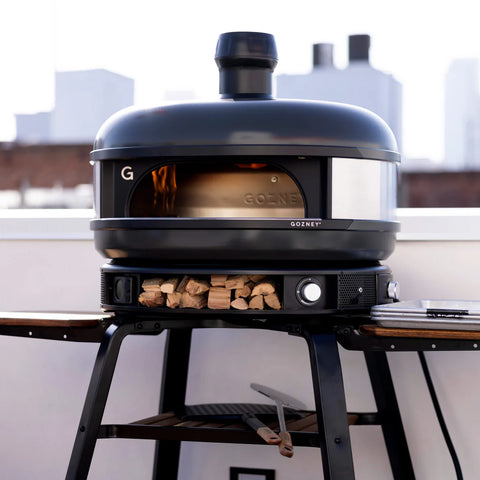 Ein eleganter Gozney Dome Pizzaofen (Multi-Fuel Gas/Holz) Off Black Bundle von Gozney steht auf einem Ständer mit gestapeltem Brennholz darunter und ist auf einem Dach mit verschwommenen Stadtgebäuden im Hintergrund platziert.