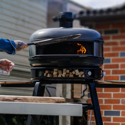 Jemand schiebt eine Pizza in den Gozney Dome Pizzaofen (Multi-Fuel Gas/Holz) Off Black Bundle von Gozney, aus dem Flammen lodern. Der Ofen steht auf einem Sockel, unter dem Holz gelagert ist, vor einer Backsteinwand.