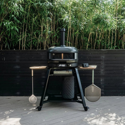 Ein Gozney Dome Gen 2 Pizzaofen - Hybrid Brennstoffsystem von Gozney mit Schornstein steht auf einer Terrasse, umgeben von Bambus und einem schwarzen Zaun. An den seitlichen Ablagen hängen Kochutensilien, und darunter ist die Radabdeckung verstaut.