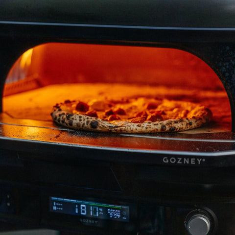 Im Inneren des Gozney Dome Gen 2 Pizzaofen - Hybrid Brennstoffsystem backt eine Pizza mit verkohlter Kruste, die orange leuchtet. Das Gozney-Bedienfeld und das Logo machen ihn zu einem auffälligen Mittelpunkt für jede Outdoor-Küche.