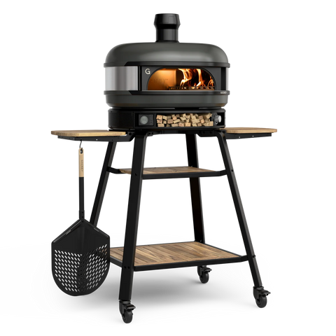 Ein Gozney Dome Pizzaofen (Multi-Fuel Gas/Holz) Off Black Bundle von Gozney wird auf einem Ständer mit Rädern und Holzregalen gezeigt, im Inneren brennt ein Feuer, an der Seite hängt ein Pizzaschieber und auf dem unteren Regal gelagertes Brennholz.