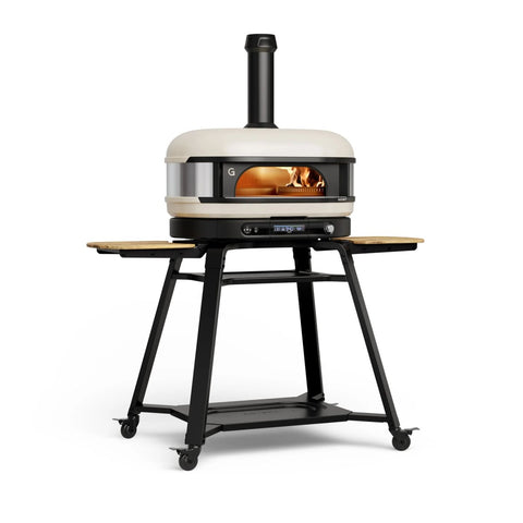 Der Gozney Dome Gen 2 XL Pizzaofen - Hybrid Brennstoffsystem von Gozney, in creme mit Schornstein, Sichtfenster und digitaler Steuerung, steht auf einem schwarzen Rollständer mit Ablageflächen. Im Inneren glüht ein Feuer, bereit für perfekte Outdoor-Pizzen.