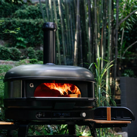 Ein schwarzer Gozney Dome Gen 2 XL Pizzaofen - Hybrid Brennstoffsystem von Gozney steht auf einem Holztisch inmitten von üppigem Grün und Bambus und schafft eine professionelle Outdoor-Küchenatmosphäre im Garten.