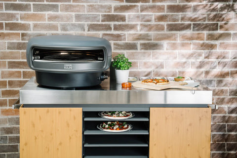 Ein Everdure KILN Gas-Pizzaofen mit rotierendem Stein steht elegant auf einem Holzschrank mit glatter Metalloberfläche. Zwei frisch gebackene Pizzen locken den eifrigen Pizzaliebhaber, ergänzt durch eine kleine Topfpflanze für Grünzeug. Die rustikale Backsteinmauer im Hintergrund trägt zu diesem ultimativen Pizzaerlebnis bei.