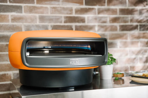 Der orange-schwarze Everdure KILN Gas-Pizzaofen mit rotierendem Stein steht auf einer Edelstahloberfläche vor einer Backsteinmauer im Hintergrund. Eine kleine grüne Pflanze ist seitlich teilweise sichtbar und ist damit perfekt für alle Pizzaliebhaber, die ein authentisches Pizzaerlebnis suchen.