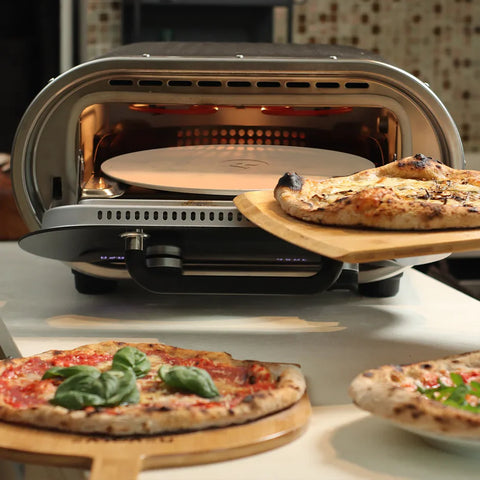 Der Evolve Smart Elektro Pizzaofen von Revolve für Innen- und Außenbereich mit rotierendem Pizzastein backt eine Pizza, während zwei fertige Pizzen - eine mit Basilikum - auf Holzschiebern vor dem Ofen liegen.