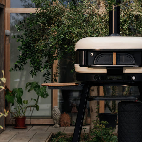 Ein Gozney Dome Gen 2 Outdoor-Pizzaofen mit Schornstein steht auf einem schwarzen Sockel in einem Garten, in der Nähe von Bäumen und Glastüren - perfekt für die Kombination mit der Gozney Pizzaofentür für Dome (Gen 2) und Ihrem Pizzaofen-Zubehör.