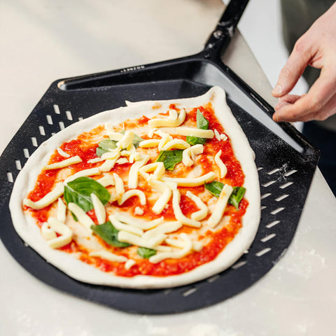 Eine Person bereitet sich darauf vor, eine tropfenförmige Pizza mithilfe eines Gozney Balance Aluminium Pizzahebers auf eine Kochfläche zu legen. Die mit Tomatensoße, Käse, Basilikumblättern und anderen Zutaten belegte Pizza ruht auf der schwarzen perforierten Schaufel, die über einen ergonomisch geformten Griff für eine einfache Handhabung verfügt.