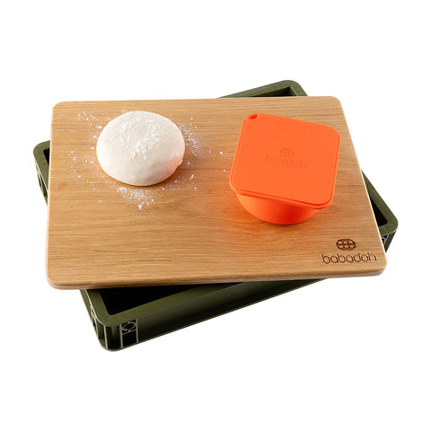 Auf einem olivgrünen, rechteckigen Tablett liegt ein Holzbrett mit einer runden, mit Mehl bestäubten Teigkugel und einer orangefarbenen Schale. Sowohl das Holzbrett als auch die orangefarbene Schale sind mit dem Logo „Babadoh“ versehen, was die Babadoh Box – Olive zu einem idealen, lebensmittelsicheren Accessoire für jeden Pizza-Liebhaber macht.
