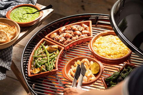 Ein runder Grill ist mit verschiedenen Tonschalen gefüllt, die gekochtes Essen enthalten. Zu den Gerichten gehören mit Speck umwickelte Speisen, ein cremiges gelbes Gericht, sautierte grüne Bohnen, Garnelen mit Zitrone und gegrillte grüne Paprika. Die keramische Pizzaofen-Grillschale FUMOSA von Fumosa sorgt für schonende Zubereitung. Pesto und Brot stehen auf einem Beistelltisch. Eine Person benutzt eine Zange am Grill.