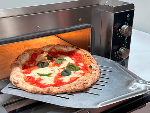 Eine heiße neapolitanische Pizza mit geschmolzenem Käse und Basilikum wird mithilfe einer Metallschaufel aus einem Resto Italia Hooven 341 Elektro Pizzaofen (Digital) gelöst, während Sorrento-Keramiksteine für eine perfekt knusprige Kruste sorgen.