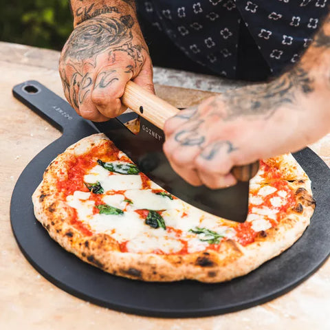 Eine Person mit Tätowierungen an beiden Händen verwendet einen gebogenen Pizzaschneider mit Holzgriff, um eine Pizza Margherita auf einem schwarzen runden Pizzastein zu schneiden. Der Gozney Pizza Servierteller (45 cm) trägt die goldene Kruste, Tomatensoße, geschmolzenen Mozzarellakäse und frische Basilikumblätter.