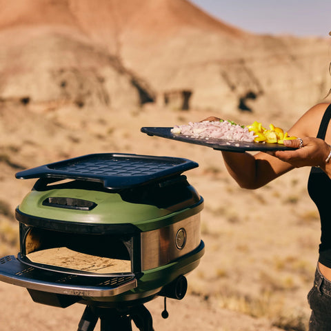 Eine Person genießt ein Outdoor-Kocherlebnis und hält ein Tablett mit geschnittenem Gemüse neben einem grünen tragbaren Ofen. In der felsigen Wüstenlandschaft liegt ein Pizzaboden im Ofen. In einem schwarzen Oberteil gekleidet, sonnen sie sich im Sonnenlicht, das die Gozney Servierplatte für Tread Roof Rack und die Szene gleichermaßen beleuchtet.