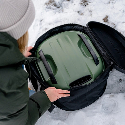 Eine Person in einer grünen Jacke und einer beigen Mütze legt ein mysteriöses grünes Objekt mit schwarzen Griffen in eine Gozney Venture Abdeckhaube & Tragetasche für Tread auf dem schneebedeckten Boden. Der Zweck des Objekts ist unklar, aber es scheint ein Behälter oder Gerät zu sein.