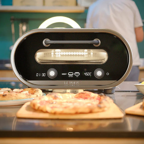 Der Evolve Smart Elektro Pizzaofen von Revolve steht auf der Küchenarbeitsplatte. Mit digitaler Anzeige für Zeit und Temperatur ist er von frisch gebackenen Pizzen auf Holzplatten umgeben.