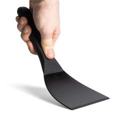 Eine Hand hält einen schwarzen Spatel mit flacher, breiter Klinge und ergonomischem Griff, mit der Klinge nach unten gerichtet. Dieser FUMOSA flexibler Teigspachtel von Fumosa ist perfekt für Pizza-Enthusiasten. Der Hintergrund ist schlicht weiß.