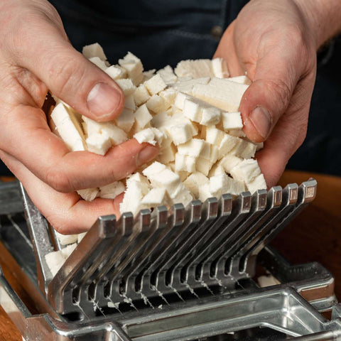 Nahaufnahme von Händen, die kleine Tofuwürfel halten, darunter ein FUMOSA Profi Mozzarellaschneider. Die Tofuwürfel sehen frisch aus, und die Hände heben sie vorsichtig hoch, sodass die Details der Tofutextur und des Fumosa-Metallschneiders sichtbar werden. Der Hintergrund ist leicht unscharf.