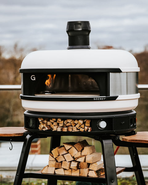Ein moderner, weißer Outdoor-Pizzaofen mit der Aufschrift „GOZNEY“ und schwarzem Schornstein, platziert auf dem Gozney Stand & Pizzaofentisch für Dome. Der Ständer verfügt über klappbare Seitentische und ein Fach darunter, das mit ordentlich gestapeltem Brennholz gefüllt ist. Der Hintergrund zeigt eine verschwommene Naturlandschaft.