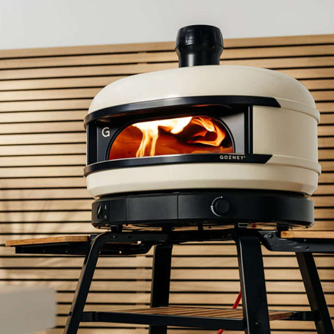 Ein Gozney Dome S1 Gas-Pizzaofen mit cremefarbener Außenseite steht auf einem schwarzen Untergestell mit seitlichen Holzablagen. Die Ofentür ist geöffnet und gibt den Blick auf ein darin brennendes Feuer frei. Der Hintergrund besteht aus horizontalen Holzpaneelen und bringt so Essen in Restaurantqualität zu Ihnen nach Hause.