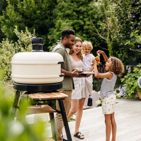 Eine vierköpfige Familie genießt einen sonnigen Tag im Freien, versammelt um ihren Gozney Dome S1 Gas-Pizzaofen. Der Vater hält ein Tablett mit Essen, die Mutter trägt ein kleines Kind und ein weiteres Kind mit lockigem Haar teilt sich ein Stück Pizza. Die Szene ist von Grün umgeben, während sie zu Hause Kochen in Restaurantqualität genießen.