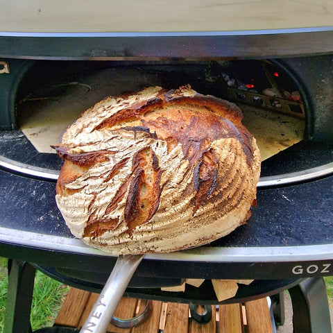 Ein frisch gebackener, rustikaler Laib Brot mit goldbrauner Kruste wird mithilfe einer Metallschaufel aus einem Pizzaofen im Freien genommen. Mit seinem handwerklichen Aussehen und den klar definierten Einschnitten ist der Laib ein Beispiel für die Finesse des Brotbackens, unterstützt durch den speziellen Gozney Steamer / Dampfinjektor für Dome Ofen Zubehör von Gozney.