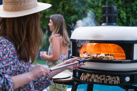 Eine Person mit Hut backt an einem sonnigen Tag Pizza in einem Gozney Gozney Dome Pizzaofen (Multi-Fuel Gas/Holz). Eine andere Person steht im Hintergrund und hält ein Getränk in der Hand. Die Szene spielt in einem Garten mit üppigem grünem Laub, was eine entspannte und angenehme Atmosphäre schafft.
