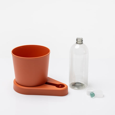 Auf einer weißen Fläche stehen eine transparente Flasche und ein roter zylindrischer Behälter mit Ausgießer. Der Behälter ruht auf einem passenden roten Ständer. Neben der Flasche sind außerdem zwei Flaschenverschlüsse und ein selbstbewässernder Kräuter-Pflanztopf (GREEVI) von Grevi zu sehen, der sich perfekt zum Anbauen frischer Kräuter eignet.
