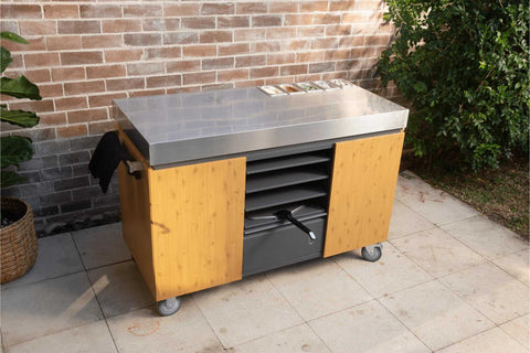 Die KILN Pizzastation von Everdure, ein hölzerner Outdoor-Küchenwagen mit einer glänzenden Edelstahl-Arbeitsfläche, schmückt die Terrasse. Sie verfügt über ein schwarzes Handtuch, mehrere Schubladen und ist von Topfpflanzen umgeben. Der Charme wird durch die Backsteinmauer und die grüne Kulisse noch verstärkt.