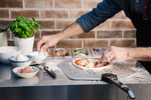 Eine Person verwendet die Everdure KILN Pizzastation, um eine Pizza zuzubereiten und verteilt Käse über Tomatensoße. In der Nähe stehen Schüsseln mit Aufschnitt und Gemüse sowie Behälter mit Belag auf einer Edelstahl-Arbeitsfläche neben einer Basilikumpflanze vor einem Backsteinhintergrund.