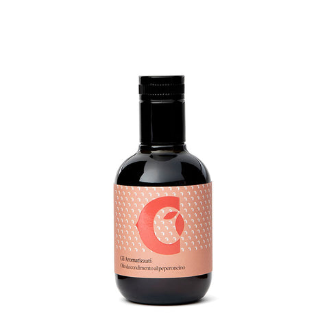 Eine kleine dunkle Glasflasche mit schwarzem Deckel ist zu sehen. Das pfirsichfarbene Etikett ist mit einem großen, stilisierten „C“ und einem abstrakten Design verziert. Der Text auf dem Etikett lautet „Olio aromatico al peperoncino“ und verrät, dass es sich um Ciccolellas extra natives Olivenöl mit Chiliaroma handelt, das in der Größe 100 ml erhältlich ist.
