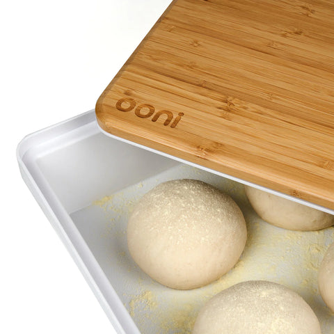 Nahaufnahme von Teigbällchen, die in einem mit Mehl bedeckten weißen Tablett liegen. Das Tablett ist teilweise mit einem Ooni Teigdeckel & Vorbereitungsbrett aus nachhaltigem Bambus bedeckt, auf dem der Markenname „Ooni“ eingraviert ist. Der Deckel ist leicht angehoben und gibt die darunter liegenden Teigbällchen frei, perfekt für die Pizzavorbereitung.