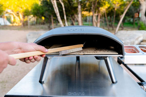 Une personne utilise une brosse pour four à pizza Ooni Ooni avec des poils métalliques pour nettoyer la surface en pierre à l'intérieur d'un four à pizza portable d'extérieur. Le four gris est posé sur une table métallique, avec des arbres et un paysage en arrière-plan flou.