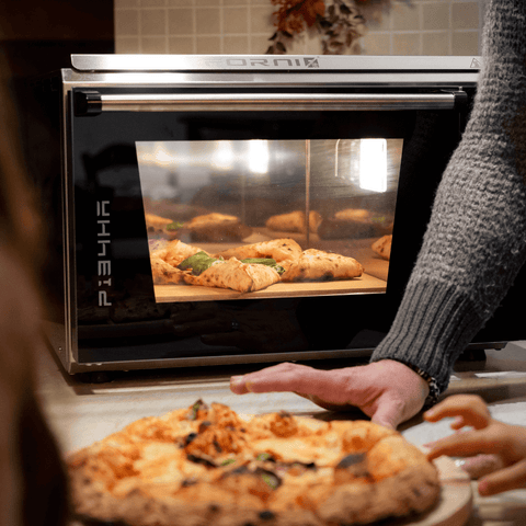 Eine Person mit grauem Ärmel ist von der Seite zu sehen und hält eine Platte mit einer frisch gebackenen neapolitanischen Pizza. Im Hintergrund wird eine weitere Pizza in einem Effeuno Pizzaofen Effeuno P134HA 509°C Evolution mit Biscotto Pizzastein gebacken. Ein Paar junger Hände streckt sich nach der Pizza auf der Platte aus.