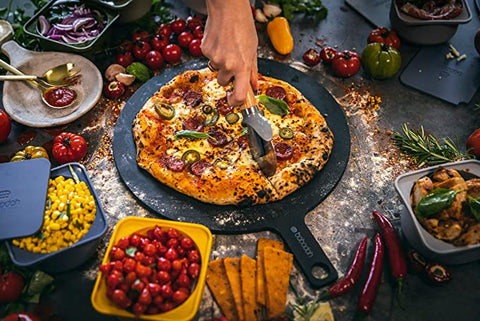 Eine Hand schneidet mit einem Pizzaschneider eine frisch gebackene Pizza, die mit Chilischoten, Peperoni und Basilikumblättern belegt ist. Rund um die Pizza auf einem Babadoh Pizza-Servierbrett aus dem Babadoh 2er SET liegen frische Zutaten wie Kirschtomaten, Mais, Zwiebeln, Chilischoten, eine Paprika und Kräuter auf einem umweltfreundlichen Holzfaserbretter-Tisch.