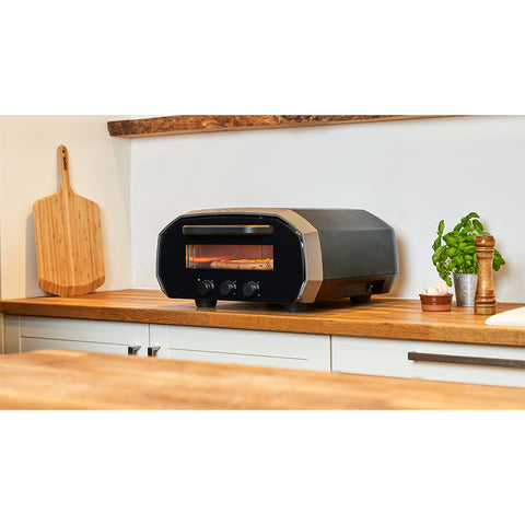 Ein Ooni Volt 12 Elektro Pizzaofen von Ooni im eleganten schwarzen Arbeitsplattendesign steht auf einer Küchentheke aus Holz. Daneben lehnt ein Pizzaschieber aus Holz an der Wand. Eine Topfpflanze und eine Pfeffermühle stehen ebenfalls auf der Theke. Der 230 V-Steckdose-Ofen backt köstliche neapolitanische Pizzen.