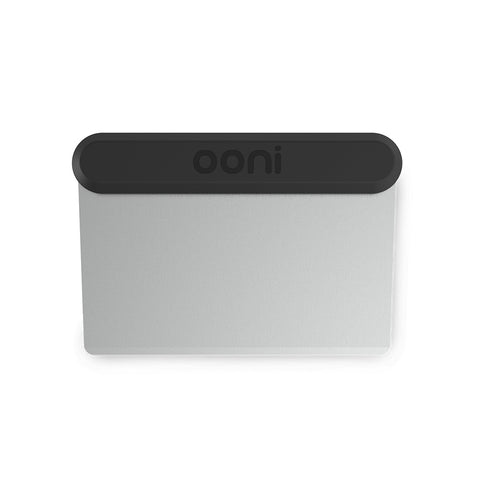 Une spatule Ooni avec une lame métallique plate et rectangulaire et un manche noir, arrondi et ergonomique. Le nom de la marque "Ooni" est gravé sur le manche. Le bord de la spatule dispose d'une arête en acier inoxydable, parfaite pour glisser sans effort sous la pâte à pizza.