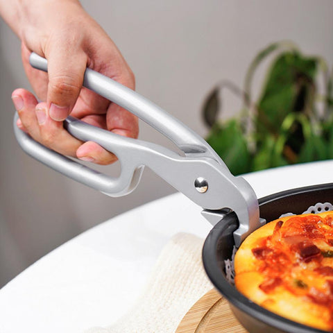 Eine Person verwendet die Pizzalovers Zange für Pizzablech, ein ergonomisch gestaltetes silbernes Greifwerkzeug, um eine heiße Pfanne mit einem Auflauf und Belag anzuheben. Die Pfanne steht auf einer weißen Oberfläche mit einer Pflanze im Hintergrund.