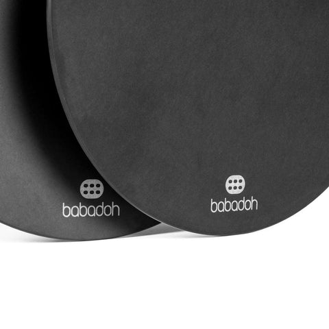 Nahaufnahme von zwei schwarzen runden Objekten mit der Aufschrift „Babadoh Pizza-Servierbrett 2er SET“ mit dem Markennamen „Babadoh“ und einem Logo mit drei Reihen von je zwei Punkten, das unten aufgedruckt ist. Die Objekte haben ein schlichtes, minimalistisches Design, ideal für die Präsentation umweltfreundlicher Pizzakreationen. Der Hintergrund ist weiß.