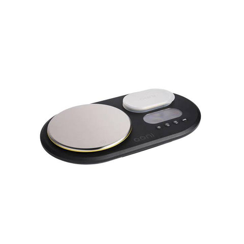 Eine schwarze Ooni Dual Digital Waage mit abgerundetem Design verfügt über zwei Edelstahl-Wiegeplattformen, perfekt für die präzise Pizzavorbereitung. Die größere runde Plattform links und die kleinere ovale rechts ergänzen einen LCD-Bildschirm und mehrere intuitive Tasten.