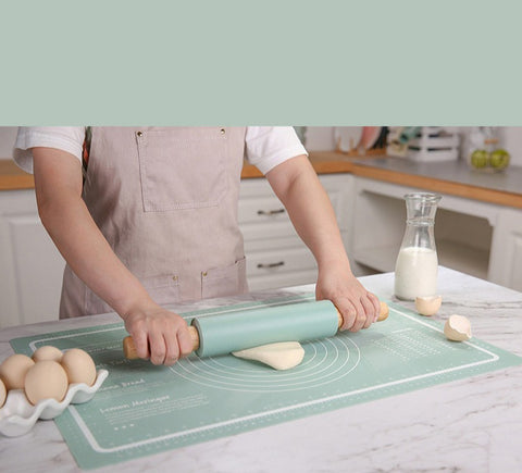 Ein Home-Pizza-Liebhaber mit einer hellen Schürze rollt in einer Küche mit einem hellgrünen Nudelholz Teig auf einer Pizzalovers Silikonmatte aus. In der Nähe stehen eine Glasflasche mit Milch und Eier in einem Eierhalter. Die Arbeitsfläche ist sauber und im Hintergrund sind Küchenutensilien zu sehen.