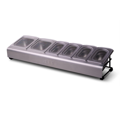 Die Ooni Pizza Topping Station ist eine elegante, moderne Servierstation für Gewürze aus silbernem Edelstahl. Sie verfügt über sechs Fächer mit durchsichtigen Kunststoffdeckeln, ideal für eine Pizzaparty. Der Markenname „Ooni“ ist deutlich sichtbar an der Seite angebracht und die Station steht elegant auf einem schwarzen Metallständer.