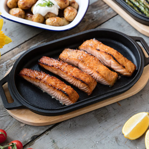 Une poêle à servir Ooni en fonte avec quatre filets de saumon grillés repose sur une planche en bois. L'extérieur du saumon est brun doré. En arrière-plan se trouve un plat de pommes de terre sautées à la crème aigre et à la ciboulette, ainsi que des asperges. Une tranche de citron et des tomates se trouvent à proximité et complètent ce bijou d'ooni sur sa base en bois.