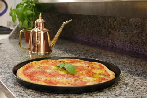 Une pizza Margherita fraîchement cuite, garnie d'une feuille de basilic, est posée sur un plan de travail en granit dans une plaque à pizza/plaque bleue (ronde) Gi.Metal noire, idéale pour les fours domestiques. À côté de la pizza se trouve un distributeur d'huile d'olive décoratif en cuivre avec un long bec verseur. Une plante de basilic se trouve à l'arrière-plan.