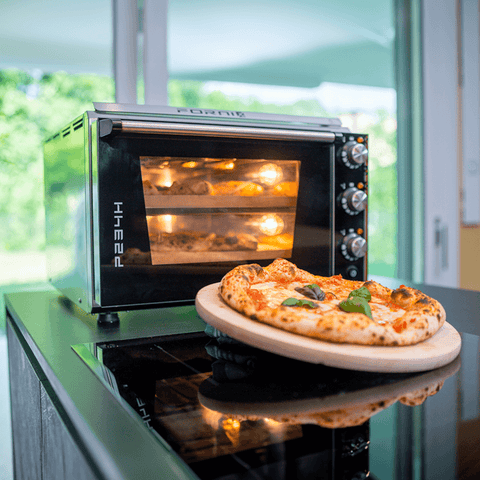 Eine frisch gebackene Pizza mit Basilikumbelag liegt auf einem Holzbrett vor einem Effeuno Pizzaofen Effeuno P234H 509°C mit 2 Backkammern. Der Ofen mit Glastür und Edelstahlknöpfen ist beleuchtet und scheint andere Biscotto-Gerichte darin zu haben. Im Hintergrund ist eine helle, sonnige Szene draußen zu sehen.